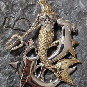 May include: A detailed pendant featuring a gold-toned merman with a crown, holding a trident. The merman is surrounded by two silver hammerhead sharks and a shark with an open mouth. A diamond is set in the merman's chest.