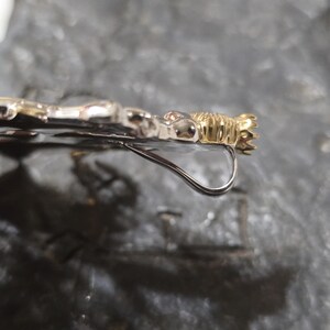 May include: Close-up of a silver and gold-toned earring. The earring features a decorative gold-colored element at the top and a curved silver hook. The design appears to be ornate and detailed.