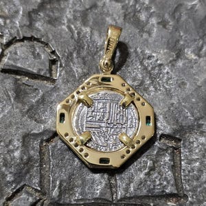 May include: A gold pendant with an octagonal frame, featuring a silver coin with intricate inscriptions. The pendant has small green gemstones and is suspended from a gold bail. The coin's design is detailed and centered within the frame.