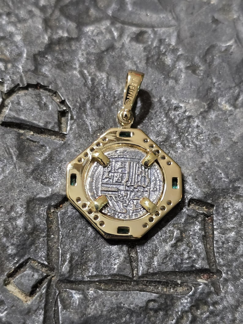 May include: A gold pendant with an octagonal frame, featuring a silver coin with intricate inscriptions. The pendant has small green gemstones and is suspended from a gold bail. The coin's design is detailed and centered within the frame.