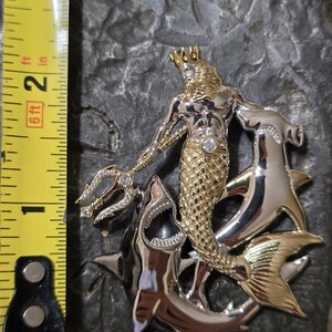 May include: A two-tone metal pendant featuring a gold-toned merman with a crown and a diamond-like gem, holding a trident. The merman is flanked by two silver-toned sharks. The pendant measures approximately 2 inches in height.