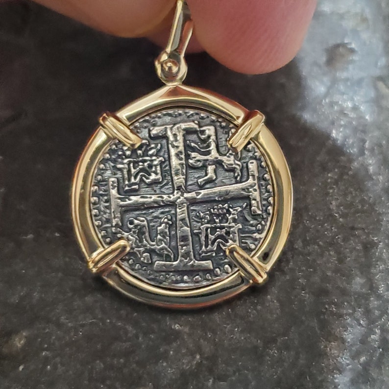 Puede incluir: Un colgante dorado con una r&eacute;plica de una antigua moneda espa&ntilde;ola con un dise&ntilde;o de cruz. La moneda est&aacute; rodeada por un marco dorado con cuatro acentos decorativos.