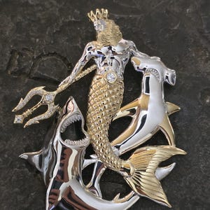 May include: A two-tone pendant featuring a merman with a crown and trident, surrounded by sharks and a hammerhead shark. The pendant is made of silver and gold-colored metal with diamond accents.