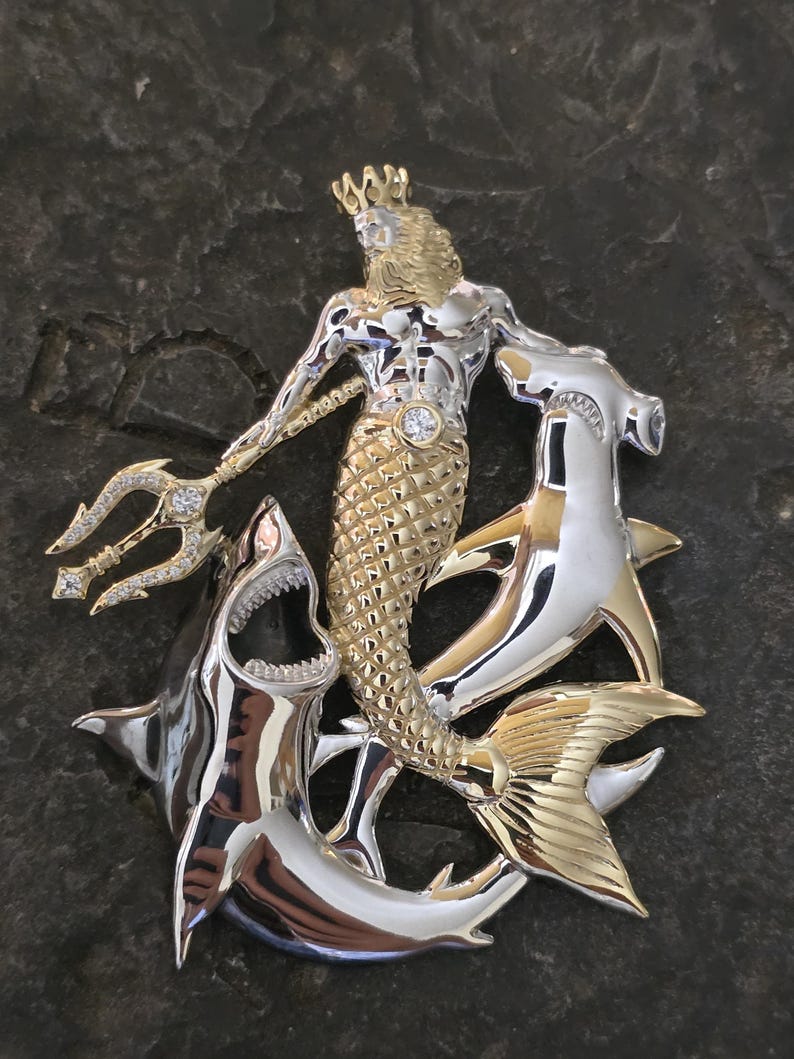 May include: A two-tone pendant featuring a merman with a crown and trident, surrounded by sharks and a hammerhead shark. The pendant is made of silver and gold-colored metal with diamond accents.