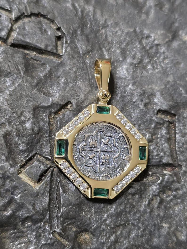 May include: A gold pendant featuring a silver coin at its center, surrounded by a gold frame. The frame is adorned with emeralds and diamonds. The pendant has a bail for attaching to a chain.