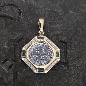 May include: A gold pendant with a silver coin center featuring a cross design. The pendant is surrounded by small diamonds and green emeralds. The pendant is octagonal in shape.