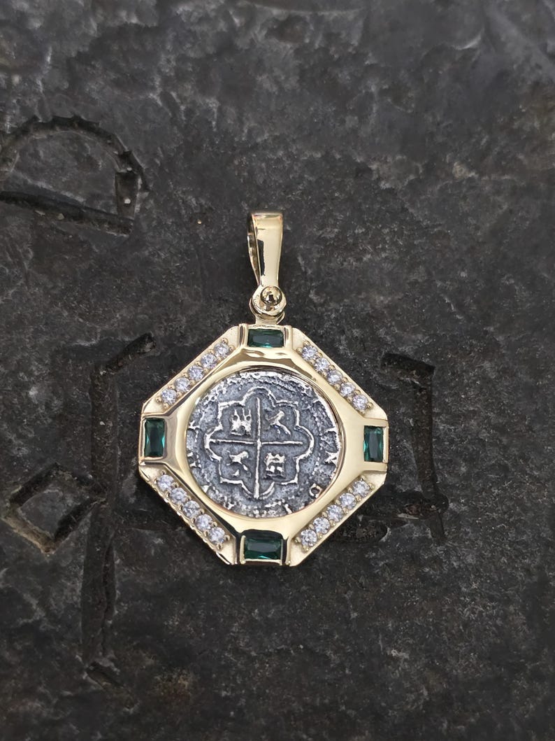 May include: A gold pendant with a silver coin center featuring a cross design. The pendant is surrounded by small diamonds and green emeralds. The pendant is octagonal in shape.