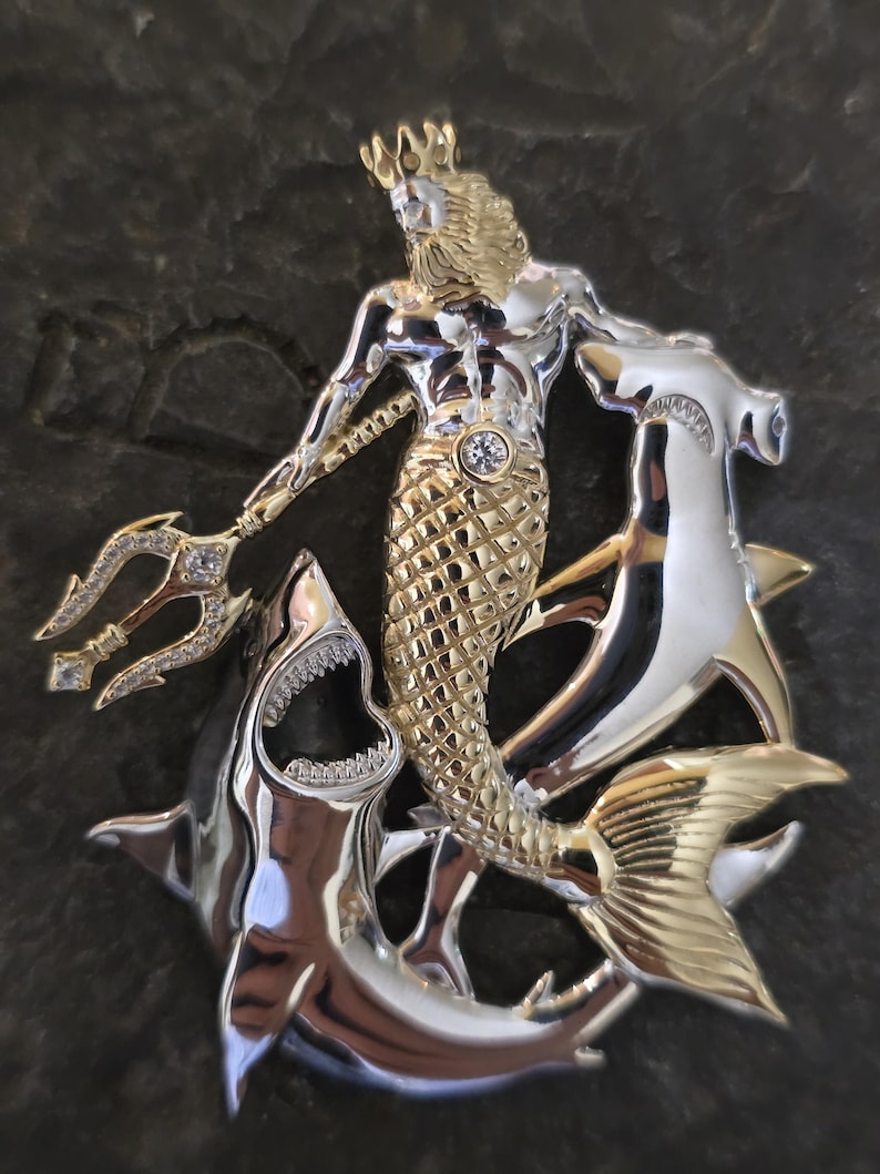 May include: A two-tone metal pendant featuring a mythical sea king with a trident, a crown, and a fish tail. The pendant is surrounded by two sharks. The king's torso is silver, and his tail and crown are gold. The sharks are silver.
