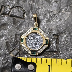 May include: A gold pendant with a silver coin center, surrounded by diamonds and emeralds. The pendant is approximately 1 inch in diameter. The coin features a cross design. The pendant is on a textured gray surface.
