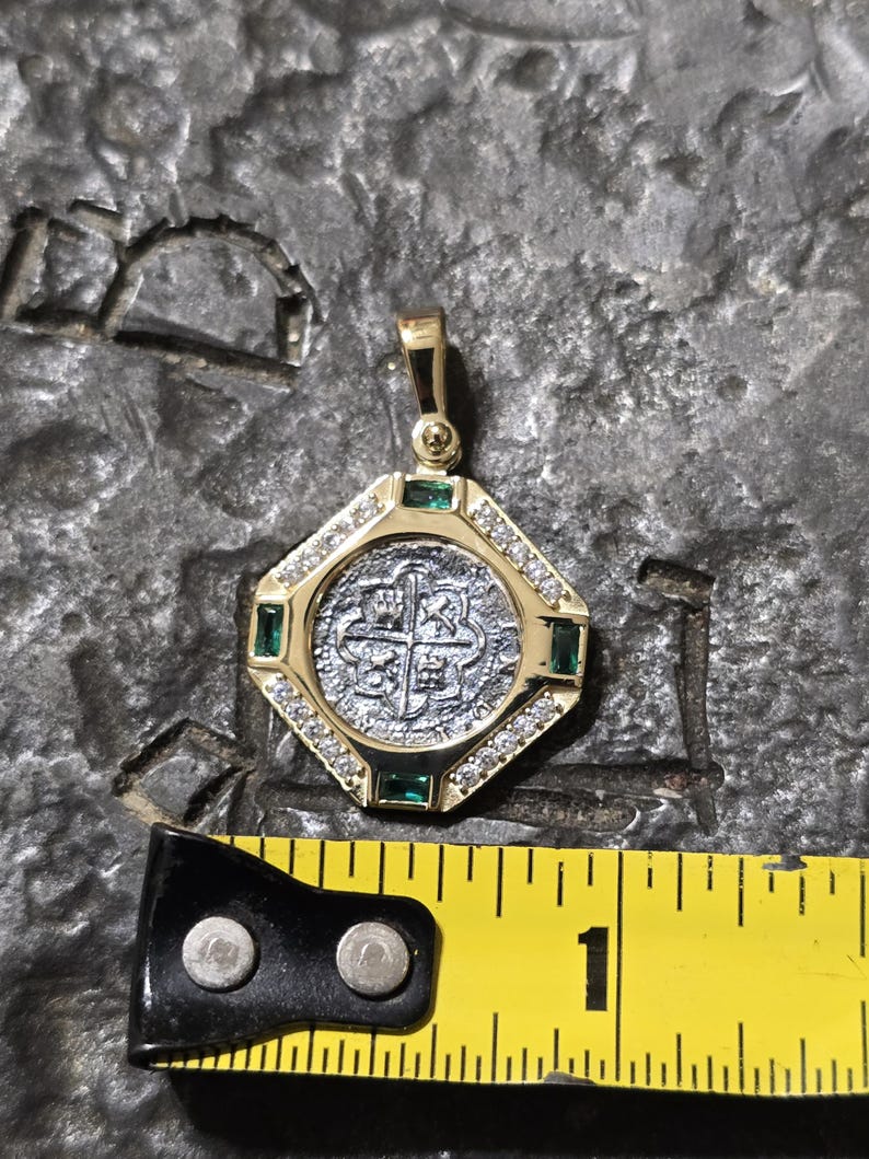 May include: A gold pendant with a silver coin center, surrounded by diamonds and emeralds. The pendant is approximately 1 inch in diameter. The coin features a cross design. The pendant is on a textured gray surface.