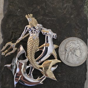 May include: A two-tone metal pendant featuring a mythical merman with a crown and trident, riding two sharks. The pendant has gold and silver tones with a diamond accent. A U.S. quarter is included for size reference.