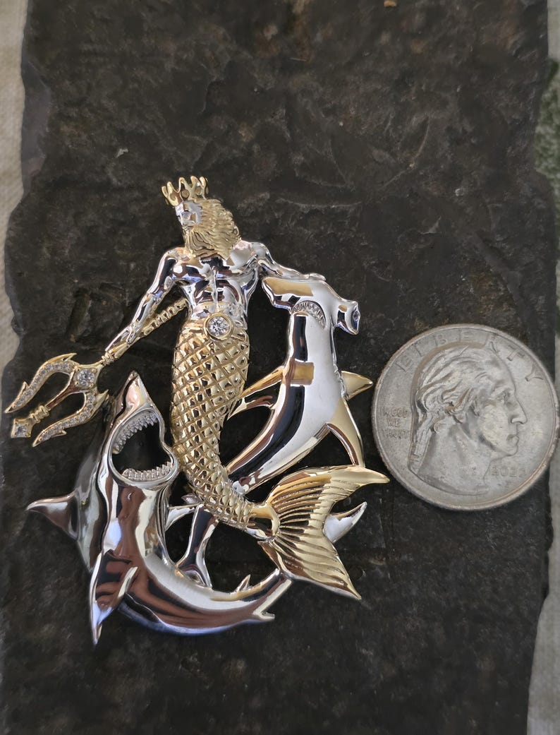 May include: A two-tone metal pendant featuring a mythical merman with a crown and trident, riding two sharks. The pendant has gold and silver tones with a diamond accent. A U.S. quarter is included for size reference.