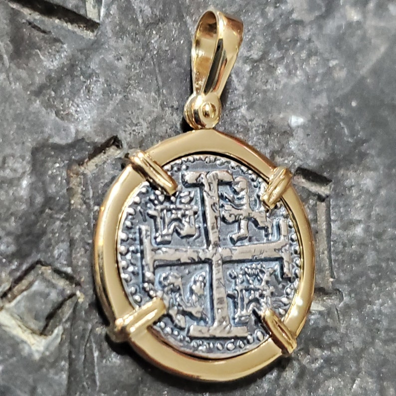 Puede incluir: Un colgante chapado en oro con una moneda de plata con un dise&ntilde;o de cruz y detalles intrincados. La moneda est&aacute; rodeada por un marco de oro y tiene un bucle en la parte superior para sujetar una cadena.