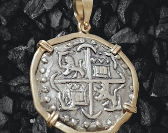 May include: A silver coin pendant with a gold frame. The coin features a cross with four shields and a ship's anchor. The coin is set in a gold frame with a bail for hanging.
