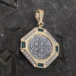 May include: A gold pendant featuring a silver coin with a cross design. The pendant is framed by a square setting with emeralds and small clear stones. The pendant has a gold bail for a chain.