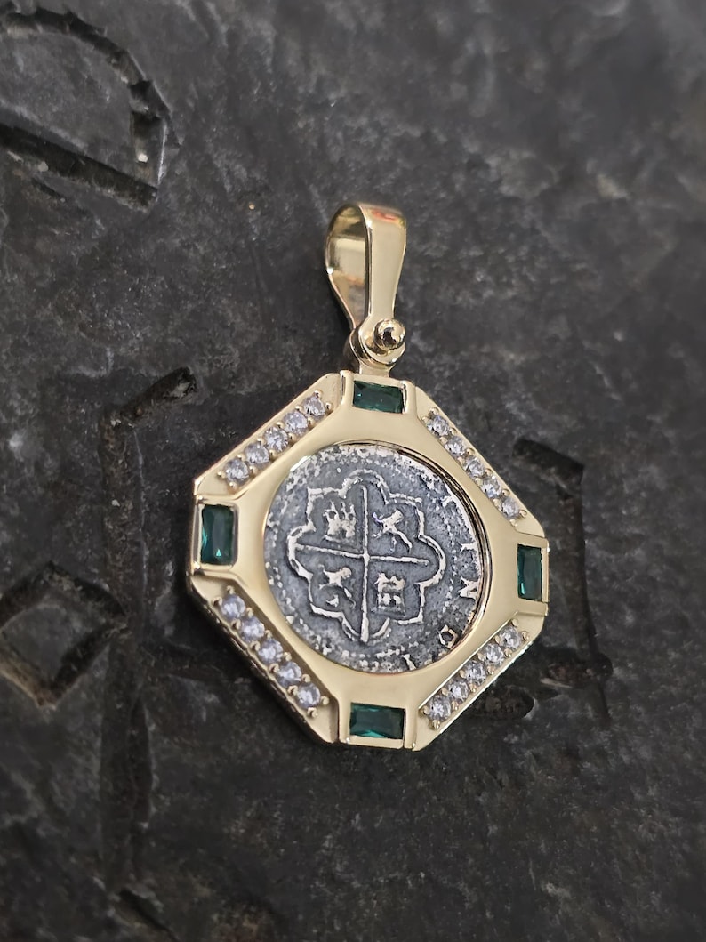May include: A gold pendant featuring a silver coin with a cross design. The pendant is framed by a square setting with emeralds and small clear stones. The pendant has a gold bail for a chain.
