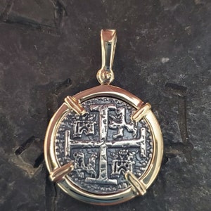 Puede incluir: Un colgante dorado con una r&eacute;plica de una moneda espa&ntilde;ola de ocho reales con una cruz y cuatro leones. La moneda est&aacute; enmarcada por un bisel dorado con cuatro barras decorativas.