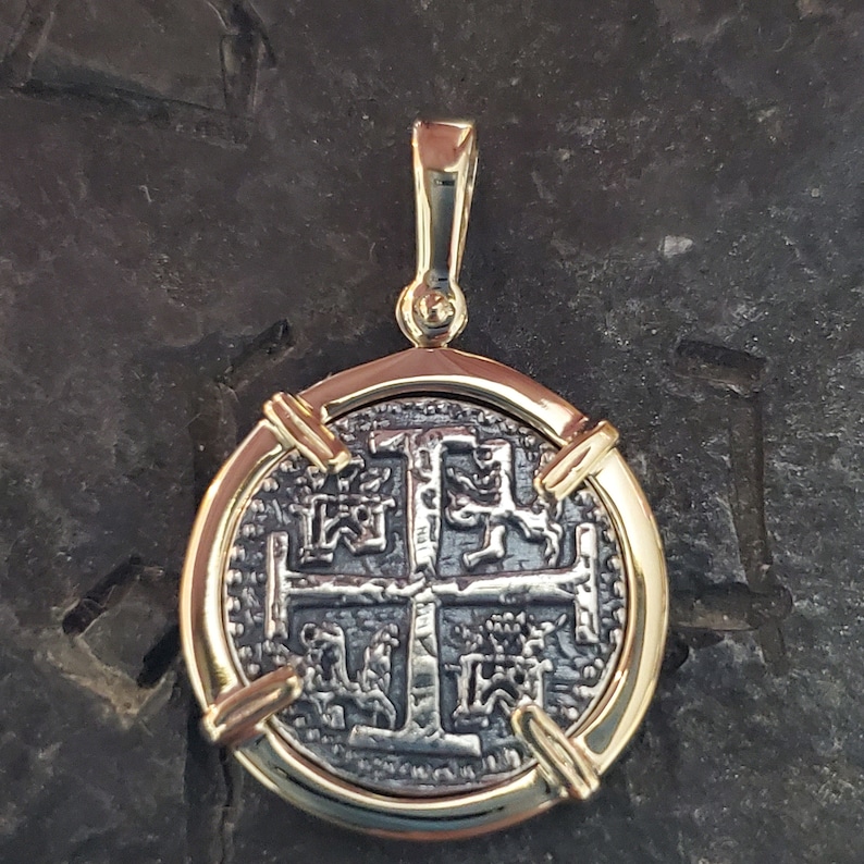 Puede incluir: Un colgante dorado con una r&eacute;plica de una moneda espa&ntilde;ola de ocho reales con una cruz y cuatro leones. La moneda est&aacute; enmarcada por un bisel dorado con cuatro barras decorativas.