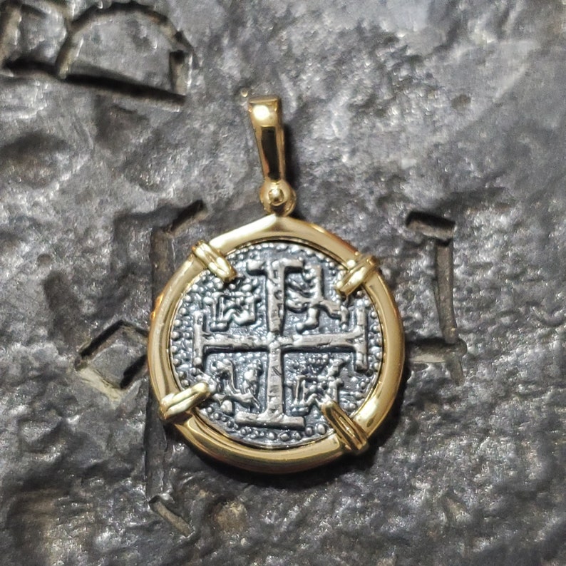 Puede incluir: Un colgante redondo de plata y oro con una cruz con detalles intrincados. El colgante est&aacute; bordeado de oro y se encuentra sobre una superficie gris texturizada.