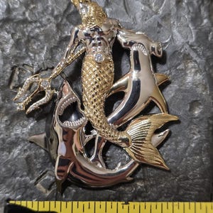May include: A two-tone metal pendant featuring a merman with a crown, holding a trident, and riding two sharks. The merman has a gold torso and tail, with a diamond-like gem on his chest. The sharks are silver.