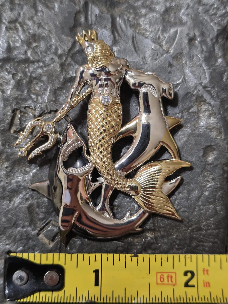 May include: A two-tone metal pendant featuring a merman with a crown, holding a trident, and riding two sharks. The merman has a gold torso and tail, with a diamond-like gem on his chest. The sharks are silver.