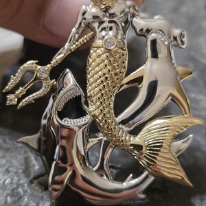 May include: A detailed pendant featuring a gold and silver merman with a crown, holding a jeweled trident. The merman is surrounded by a shark and a hammerhead shark, with intricate detailing and sparkling accents.