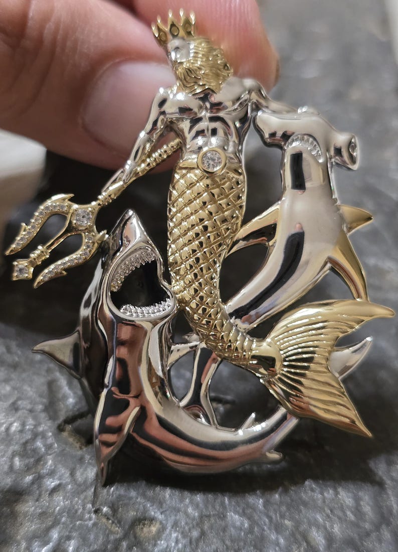 May include: A detailed pendant featuring a gold and silver merman with a crown, holding a jeweled trident. The merman is surrounded by a shark and a hammerhead shark, with intricate detailing and sparkling accents.