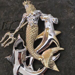 May include: A two-tone gold and silver pendant featuring a mermaid with a crown, holding a jeweled trident, and surrounded by two sharks. The mermaid's tail is gold, and the sharks are silver.