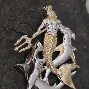 May include: A two-tone metal pendant featuring a merman with a trident, a crown, and a fish tail, flanked by two sharks. The merman is gold-colored, and the sharks are silver-colored. The pendant has a detailed design with a gemstone on the merman's chest.