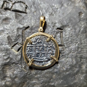 Puede incluir: Un colgante de oro con una r&eacute;plica de plata de una moneda espa&ntilde;ola de 2 reales con la inscripci&oacute;n "P.V.A." y el a&ntilde;o "1728".