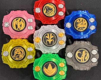 May include: A collection of seven circular, gear-shaped plastic devices in red, pink, yellow, white, green, blue, and gray. Each device features a central black circular emblem with a gold-colored animal or symbol relief and four small buttons.