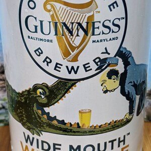 May include: A white Guinness Wide Mouth White Ale beer can. The can features a gold harp logo and the text "Open Gate Brewery Baltimore Maryland". A cartoon alligator and man are on the can. The text "Wide Mouth White Ale" is also visible.