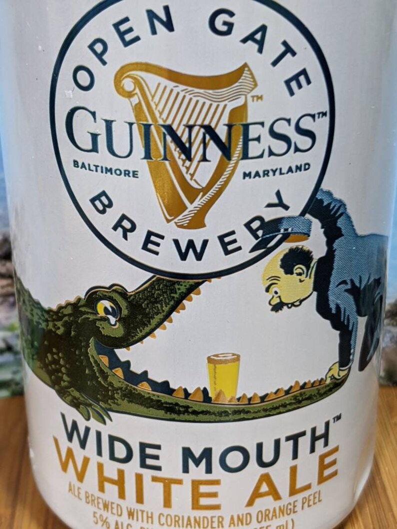 May include: A white Guinness Wide Mouth White Ale beer can. The can features a gold harp logo and the text "Open Gate Brewery Baltimore Maryland". A cartoon alligator and man are on the can. The text "Wide Mouth White Ale" is also visible.