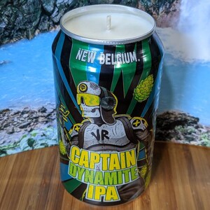 May include: A Captain Dynamite IPA candle in a repurposed beer can. The can features a green, black, and blue design with a skeleton character wearing VR goggles. The candle has a white wax top and a single wick.