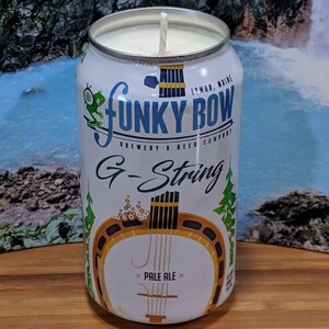 May include: A white candle in a beer can with the text "FUNKY BOW" and "G-String Pale Ale". The can features a frog graphic and a banjo illustration. The candle has a white wax and a single wick.