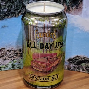 May include: A candle in a repurposed beer can. The can features the "Founders All Day IPA" label with a forest scene and a vintage station wagon. The candle has a white wax and a single wick.