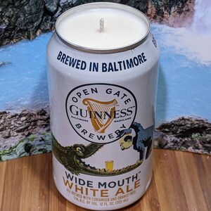 May include: A white candle in a repurposed Guinness beer can. The can features the text "BREWED IN BALTIMORE" and "OPEN GATE GUINNESS BREWER." The can also has a graphic of an alligator and a person. The candle is made with white wax.