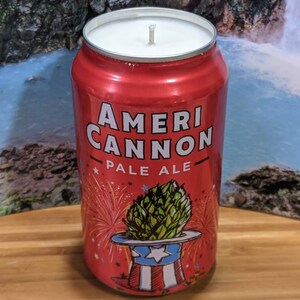 May include: A red candle in the shape of a beer can, labeled "AmeriCannon Pale Ale." The can features a patriotic design with a hop in a Uncle Sam hat and fireworks. The candle has a white wax top and a wick.