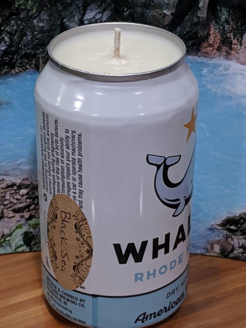 May include: A white candle in a can with a whale illustration and the words "WHALE RHODE" in black. The can also has the words "Black Sea" on a brown label and "American" in blue.