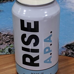 May include: White candle in a can with the words "RISE A.P.A." in black and light blue. The bottom of the can reads "MADE IN Rhode Island". The candle has a white wax and a single wick.