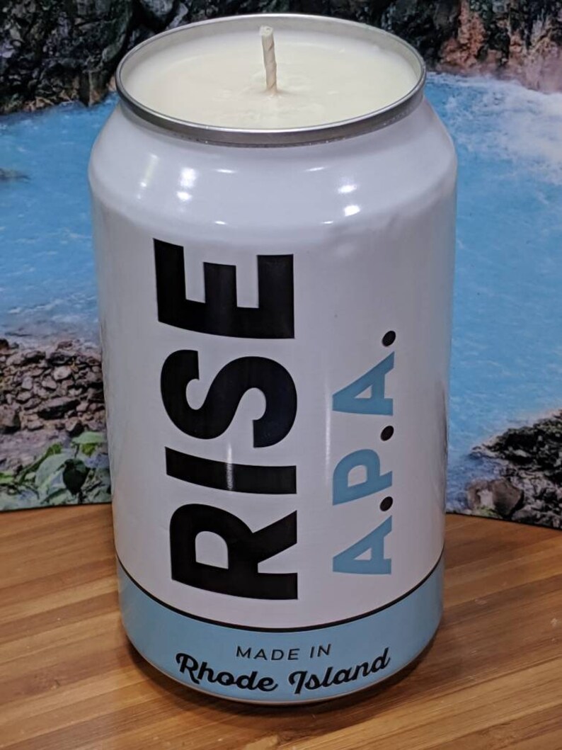 May include: White candle in a can with the words "RISE A.P.A." in black and light blue. The bottom of the can reads "MADE IN Rhode Island". The candle has a white wax and a single wick.