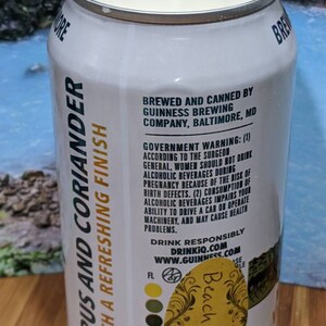May include: A white candle in a repurposed Guinness beer can. The can features text that reads "Citrus and Coriander with a Refreshing Finish" and "Beach Linen". The candle has a wick and a light-colored wax.