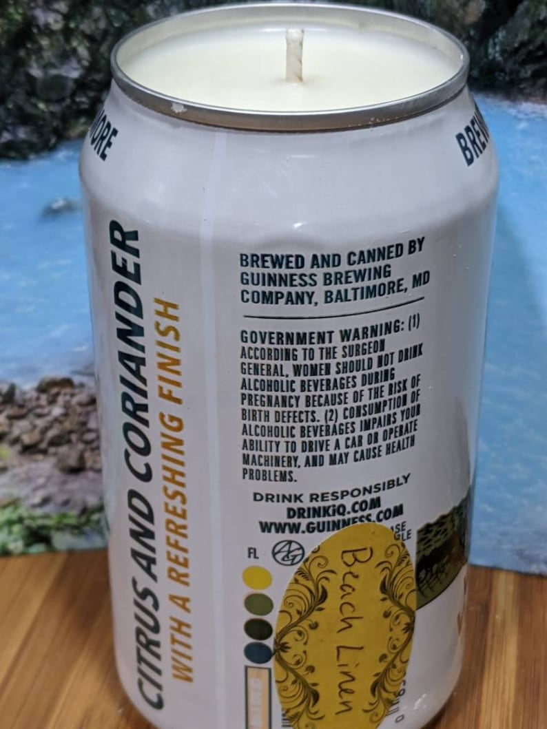 May include: A white candle in a repurposed Guinness beer can. The can features text that reads "Citrus and Coriander with a Refreshing Finish" and "Beach Linen". The candle has a wick and a light-colored wax.