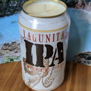 May include: A white Lagunitas IPA beer can repurposed as a candle holder. The can features the Lagunitas IPA logo in brown and gold. The top of the can is trimmed with gold and filled with a cream-colored candle.