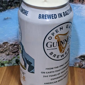 May include: A white candle in a repurposed Guinness beer can. The can features the Guinness logo, text that reads "BREWED IN BALTIMORE", and other text. The candle has a cream-colored wax and a central wick.