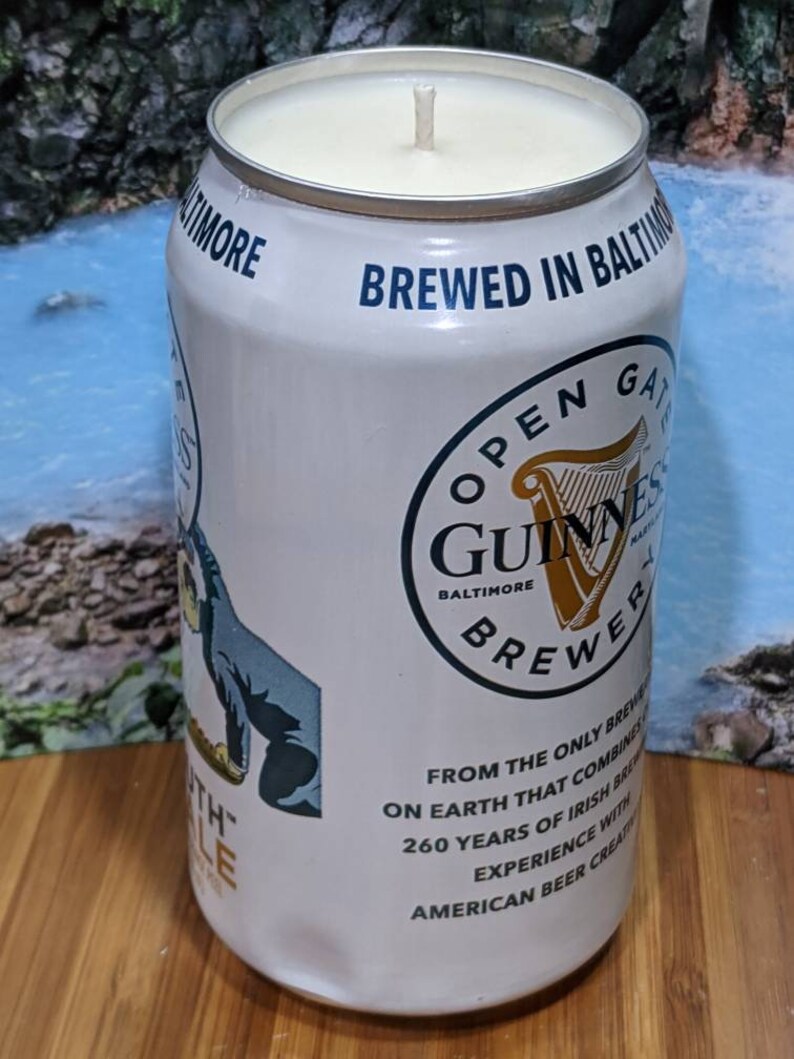 May include: A white candle in a repurposed Guinness beer can. The can features the Guinness logo, text that reads "BREWED IN BALTIMORE", and other text. The candle has a cream-colored wax and a central wick.