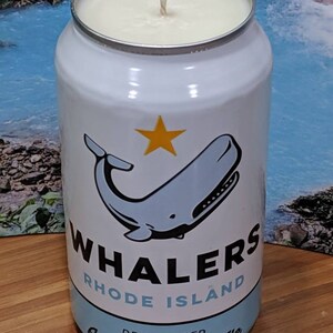 May include: A white candle in a can with a whale illustration and the text "WHALERS RHODE ISLAND American Pale Ale". The candle has a cream-colored wax and a wick.