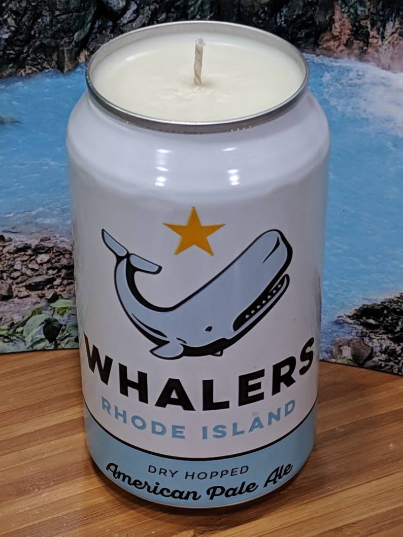 May include: A white candle in a can with a whale illustration and the text "WHALERS RHODE ISLAND American Pale Ale". The candle has a cream-colored wax and a wick.