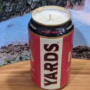 May include: A red and white candle in the shape of a beer can, with the text "YARDS" and "EST 1994". The candle has a white wax top and a wick. The can is sitting on a wooden surface.