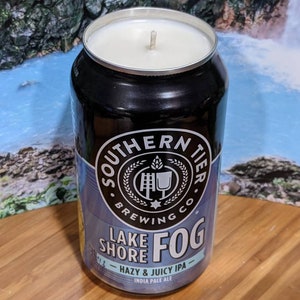 May include: A candle in a repurposed beer can from Southern Tier Brewing Co. The can is black with blue and white labels, featuring the text "Lake Shore Fog - Hazy & Juicy IPA". The candle has a white wax and a single wick.